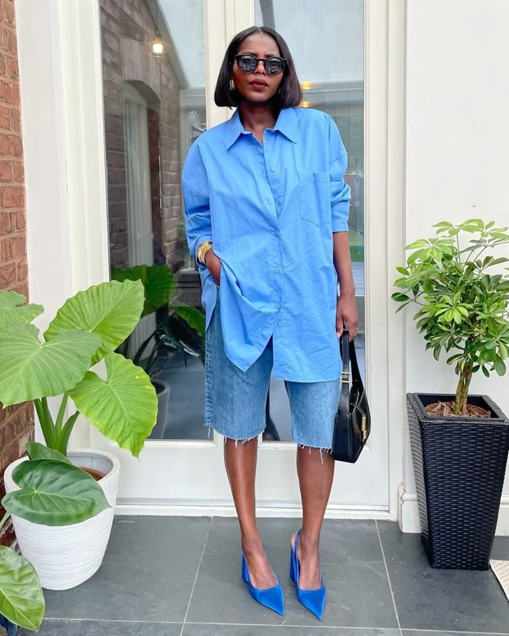 A person wearing an oversized blue button-down shirt, denim shorts, and blue high-heeled shoes, standing in a stylish indoor setting with plants.