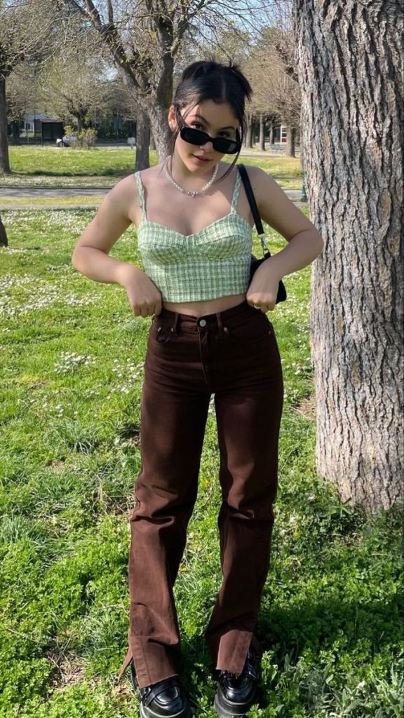 A young woman standing outdoors, wearing a green checkered crop top and brown flared pants, with sunglasses and a small handbag. She poses in front of trees and grass.