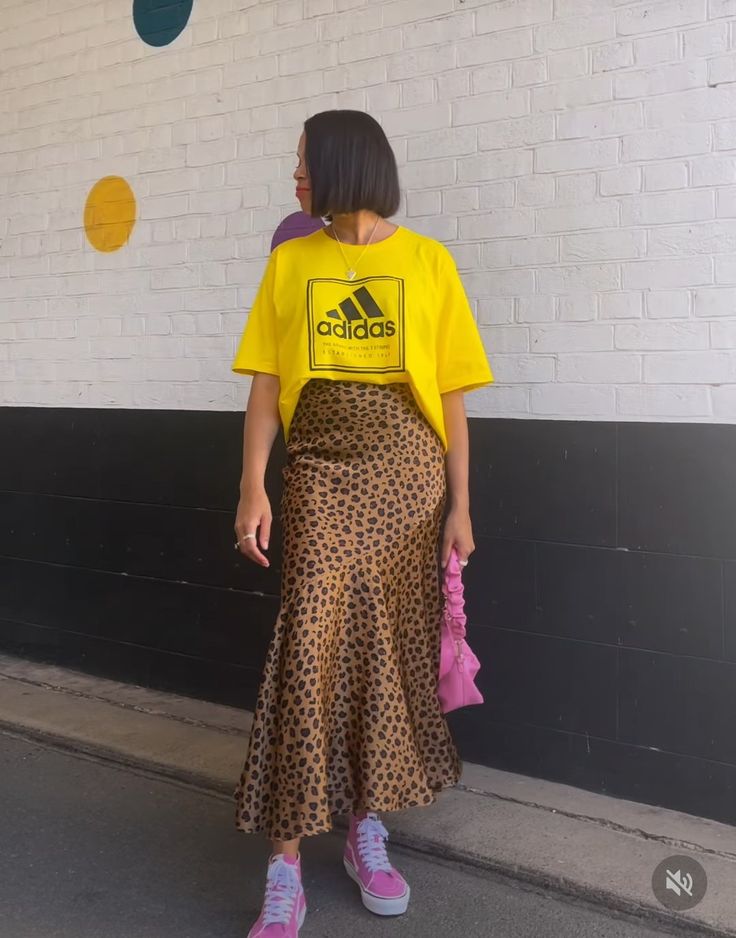 A woman wearing a yellow oversized Adidas t-shirt paired with a leopard print skirt and pink sneakers, standing in front of a colorful wall.