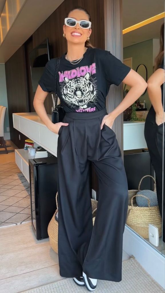 A person smiling while wearing a black graphic tee with a tiger design, high-waisted black wide-leg pants, and sunglasses, standing in a bright indoor setting.