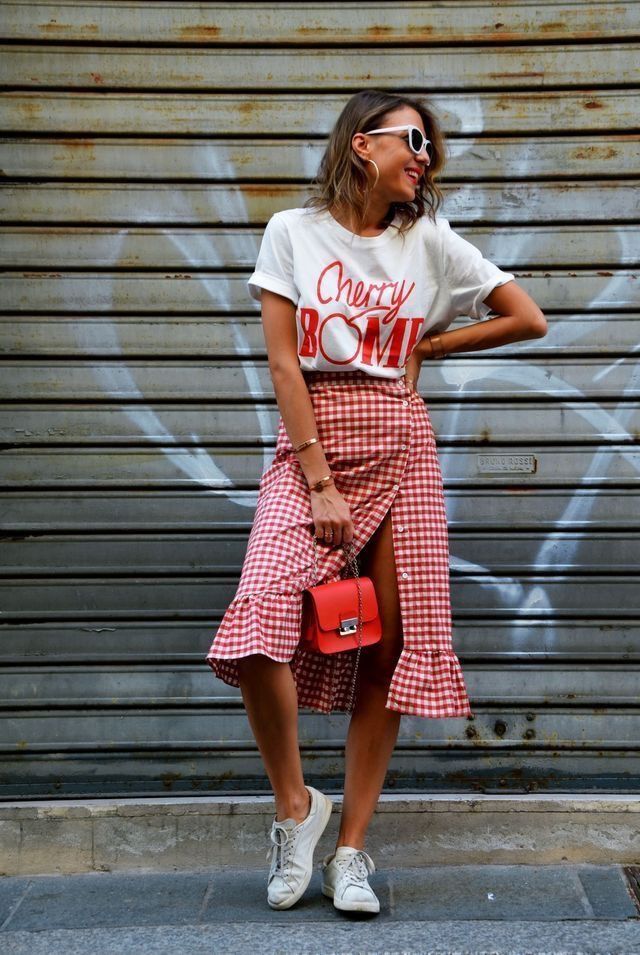 A woman stands against a metal wall wearing a white graphic t-shirt with the text 'Cherry Bomb', a red gingham skirt with a ruffled hem, and white sneakers. She accessorizes with large hoop earrings and sunglasses, holding a small red bag.