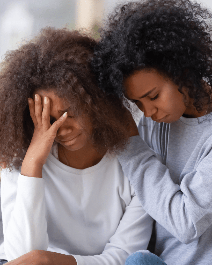 Two women embrace in a moment of support, one with her face in her hand displaying distress, while the other leans in closely providing comfort.