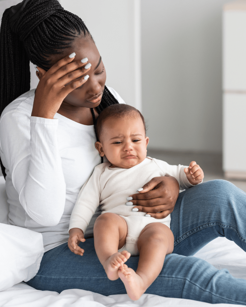 A mother sitting on a bed with a distressed baby in her arms, looking contemplative and concerned.