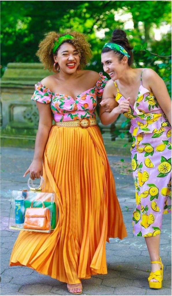 Two women smiling and walking together in vibrant summer outfits, one in a floral top and pleated orange skirt, and the other in a lemon print dress with yellow heels, both wearing colorful headbands.