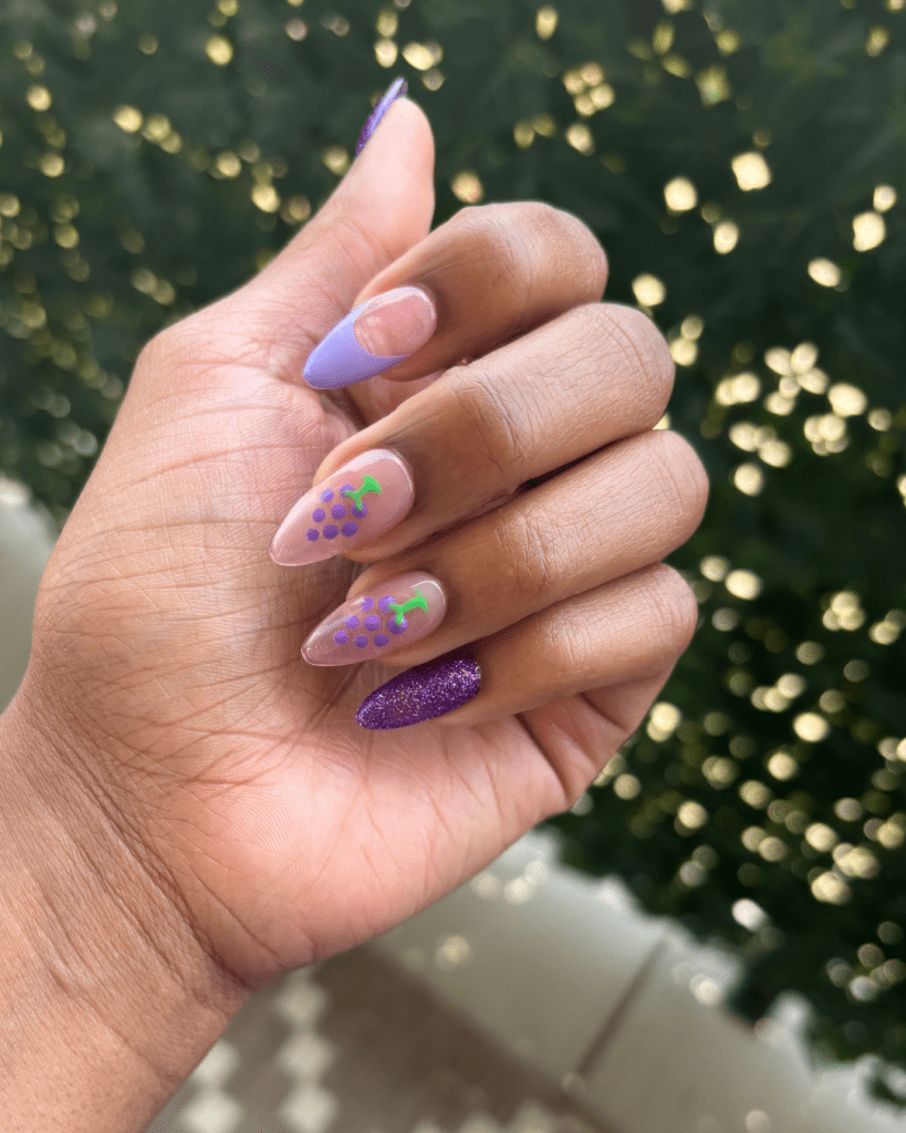 A close-up view of a hand with colorful DIY nail art featuring grape designs on a few nails, a purple glitter accent nail, and a light pink base on other nails, set against a blurred green background.