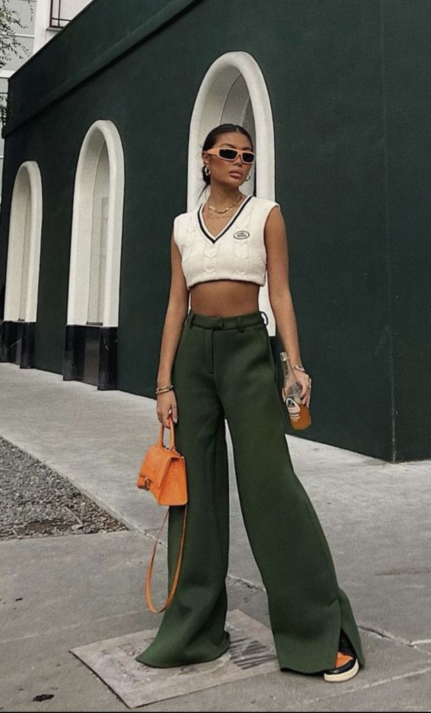 A fashionable young woman stands confidently on the street, wearing a cropped white top and wide-leg green pants. She accessorizes with orange sunglasses and a small orange handbag, showcasing a modern and playful style against a backdrop of arched green walls.
