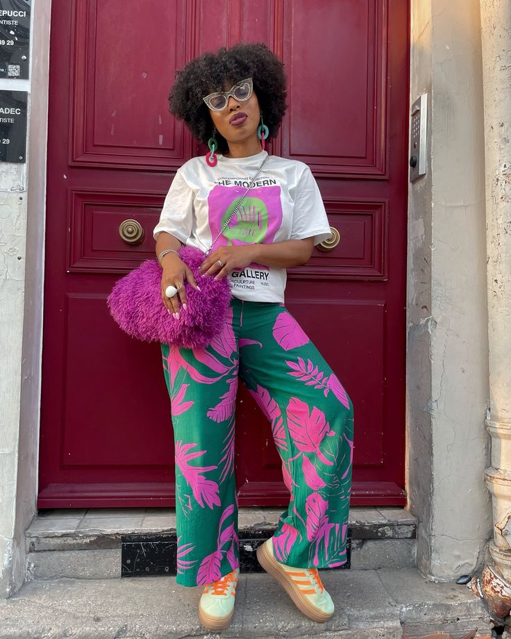 A person standing in front of a red door wearing a graphic t-shirt, bold printed pants, and chunky sneakers, holding a fluffy purple purse.