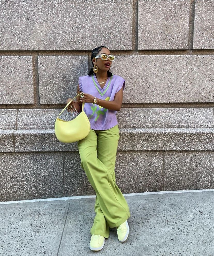 A fashionable individual stands against a textured wall, wearing a purple knitted top with a green design and light green pants. They are holding a yellow bag and wearing sunglasses, exuding a playful summer vibe.