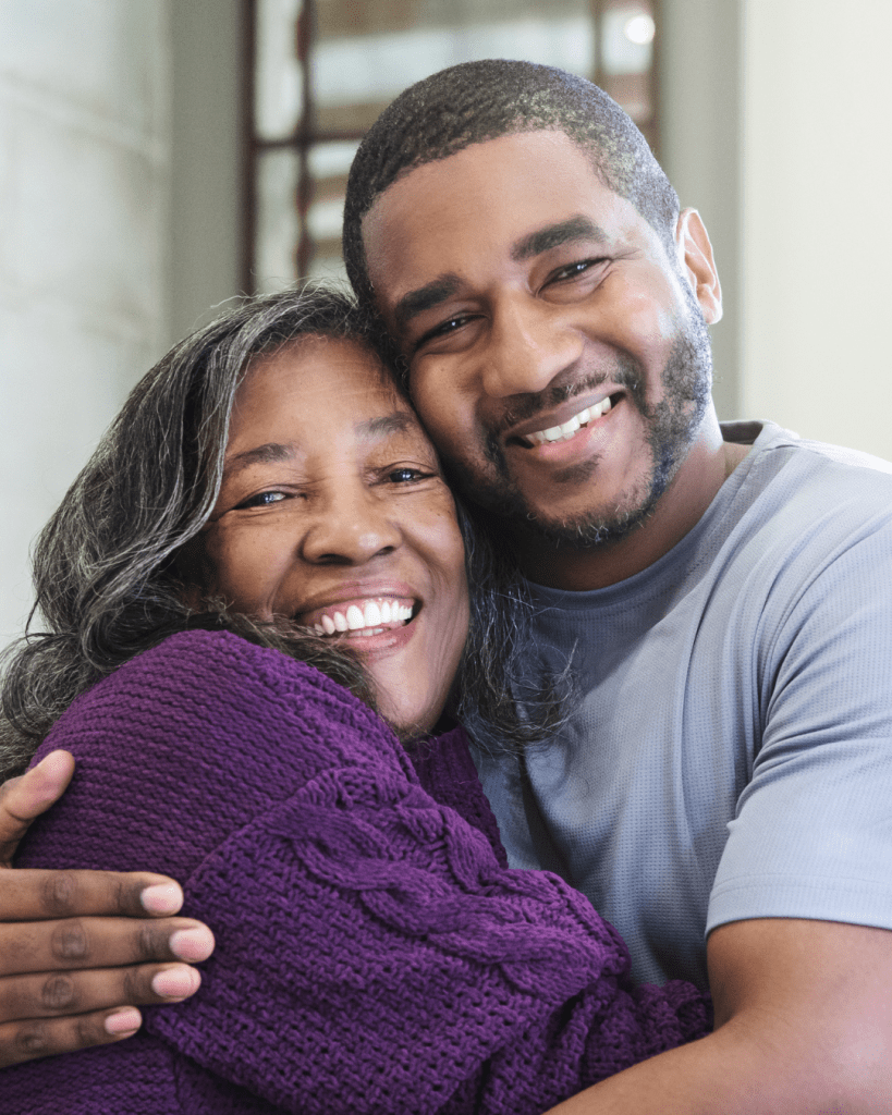 A joyful elderly woman with gray hair wearing a purple sweater smiles warmly while embracing a young man with a beard, who is wearing a gray shirt. They seem to share a moment of love and connection.