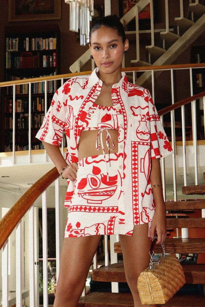 A person wearing a matching red and white patterned crop top and skirt, standing in a home interior with wooden stairs and bookshelves in the background.