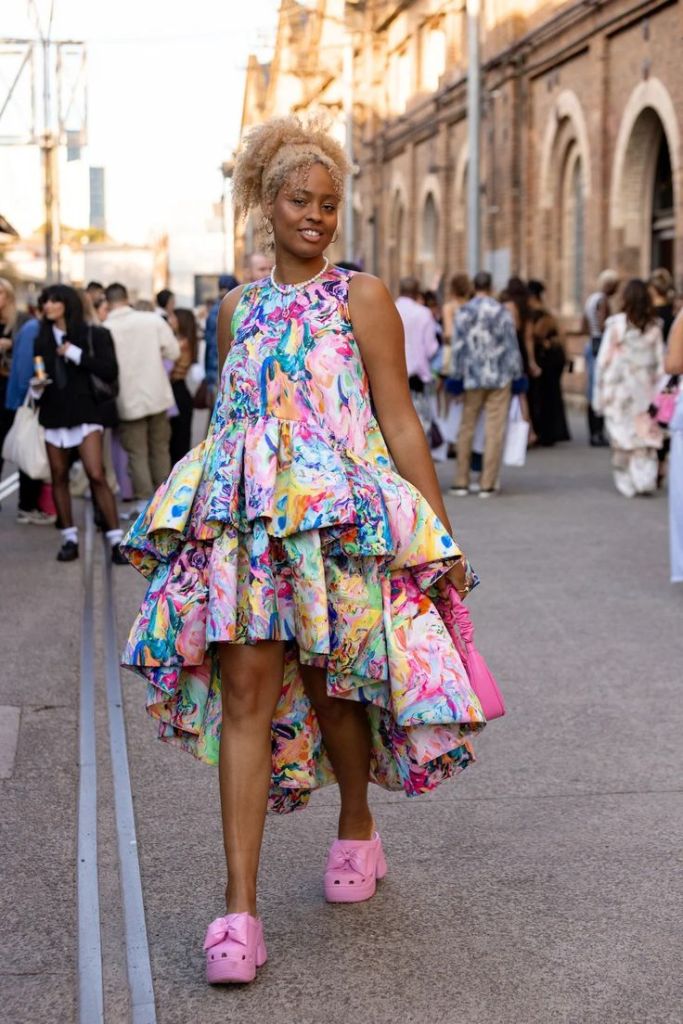 A person walking in a vibrant, multicolored dress with ruffled layers, styled with pink crocs, in an outdoor urban setting.