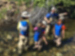 3 children in waders standing in the water watching a guide tell them about a fly fishing rod