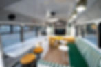 Interior of a custom skoolie conversion featuring a modern lounge area. The space includes a white marble-look countertop with mustard yellow stools, a built-in TV, two curved mustard-colored armchairs, a green and white striped cushioned bench, and small round tables. The design incorporates white paneling with green accents, large windows, and warm wood flooring."