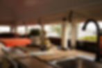 Cozy skoolie interior with a bed covered in an orange comforter, positioned next to large panoramic windows. A small indoor garden setup with a potted plant, a jar of tea, and a rustic mug sits on the wooden countertop.