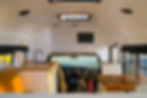 Front interior of a skoolie conversion with a view of the driver's cabin. A modern kitchenette with wood countertops, a black faucet, and white cabinetry is positioned near the entrance. A rustic wooden dining table and a fire extinguisher are also visible.