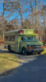A custom-built skoolie conversion on a Chevy Express chassis, painted in matte olive green. The bus is parked outdoors, reflecting trees on its large side windows. The front showcases a black grille with a gold Chevrolet emblem.