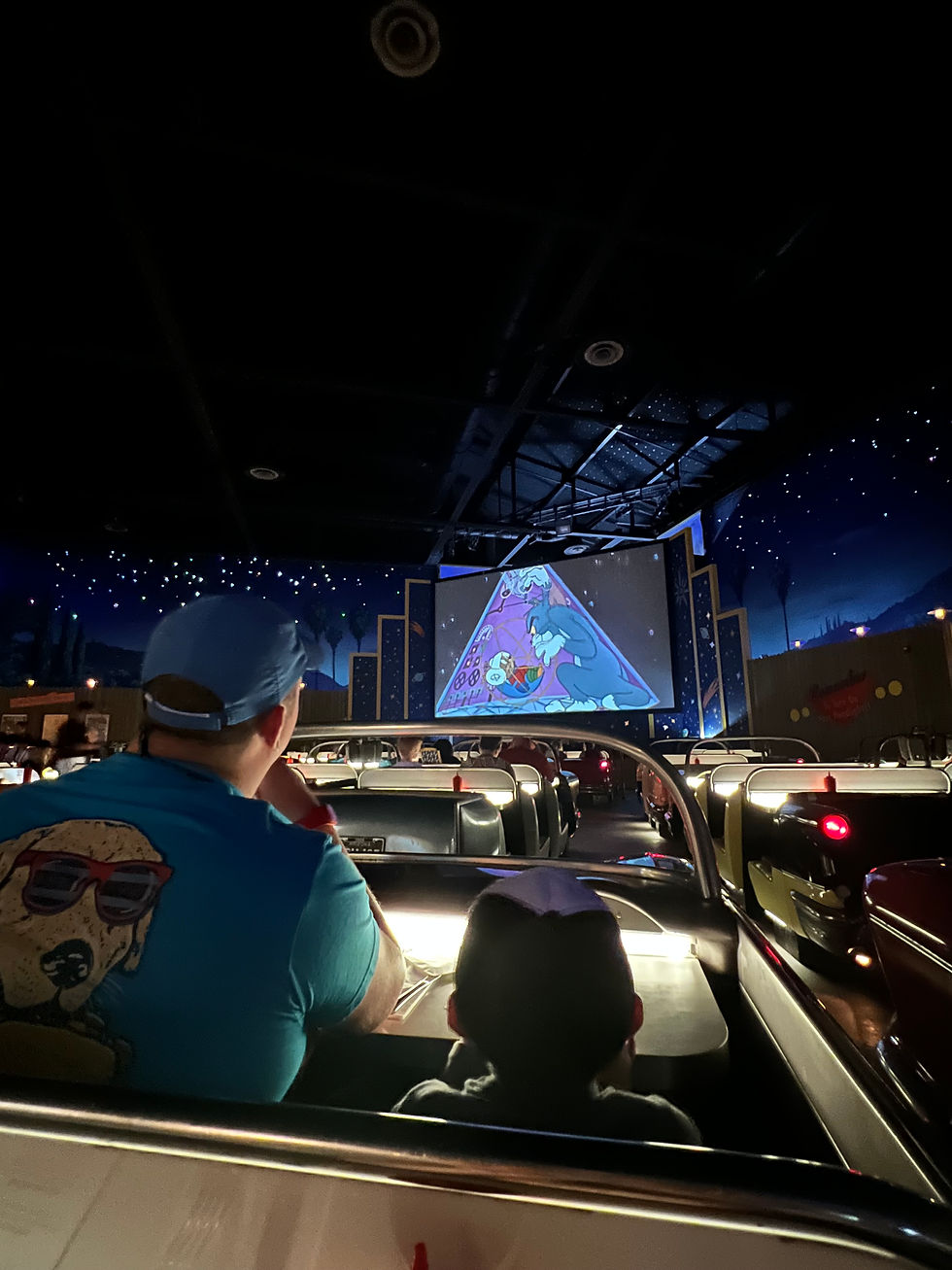 Drive in diner at Disney World man and child sitting in a car waiting for food watching cartoons on a large projector screen with stars in the sky and other cars with other people.