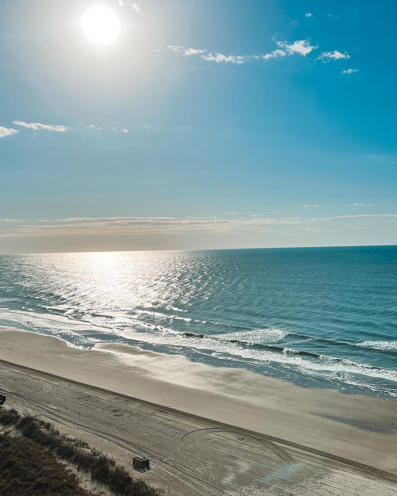 Sun-soaked beach with blue sky, a few clouds, gray sand and shrubs.