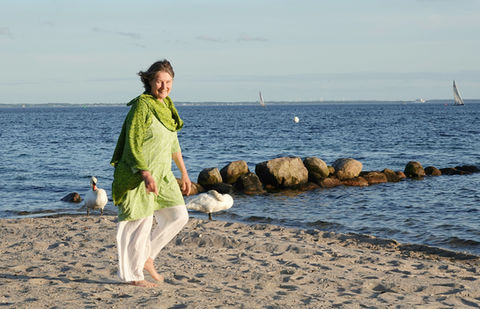 Frau am Strand mit grünem Oberteil, weißer Hose, See und Segelboot. elkemariestoehr