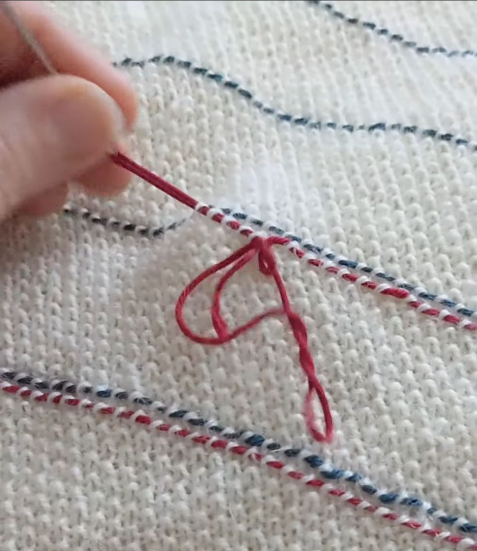 a woman's hand darning a red thread of yarn underneath a green line of yarn on a jumper