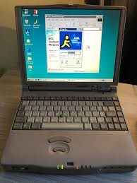 Vintage laptop with a beige color on a desk, screen displaying AOL Instant Messenger. Desktop icons on teal background, nostalgic tech vibe.