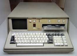 Grey vintage computer with a small screen, keyboard, and dual tape drives. The IBM logo appears on the front, set against a neutral background.