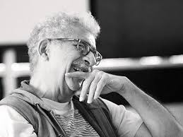 Elderly man with glasses and curly hair, smiling, hand on chin. Black and white setting. Relaxed, thoughtful mood.