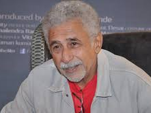 Older man with gray hair and beard wearing a red shirt and gray jacket, smiling slightly. Dark, text-covered background. Calm mood.