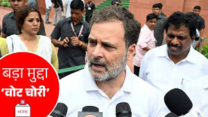 A man in a white shirt speaks to microphones outside, surrounded by others. Red sign with Hindi text and a vote symbol is visible.