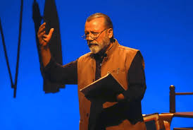 A man in a brown vest gestures passionately while reading from a book against a blue background, suggesting a dramatic or insightful mood.
