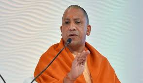 Man in orange robe speaks into a microphone against a white textured background. His expression is calm and focused.