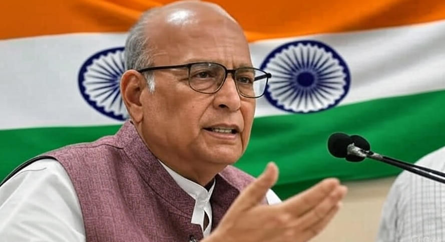Elderly man in glasses speaks passionately at a microphone. Indian flag in background. Wearing a maroon vest, conveying a serious mood.