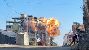 Explosion engulfs a building on an urban street under clear blue sky. A person on a bicycle observes from the sidewalk. Tense atmosphere.