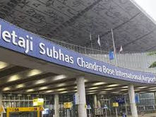 Sign for Netaji Subhas Chandra Bose International Airport on a modern building. Overcast sky and green foliage are visible in the background.