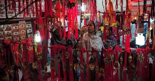 Women in a vibrant market browse red and gold decorative items under bright lights. The mood is festive and lively.