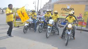Five motorcyclists in yellow shirts and helmets line up on a street. A man waves a yellow flag. "Rapido" branding visible in the background.