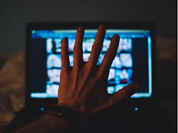 A hand covers a laptop screen displaying blurred images. The setting is dimly lit, creating a mysterious mood.