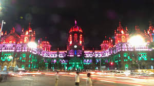 Historic building illuminated with vibrant red, purple, and green lights at night. Blurred cars pass by, and people are in foreground. Energetic mood.
