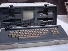 Vintage portable computer with a built-in screen and keyboard in a hard case. The device is gray and beige, on a white surface.
