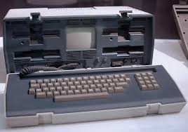 Vintage portable computer with a built-in screen and keyboard in a hard case. The device is gray and beige, on a white surface.