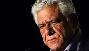 Elderly man with gray hair and serious expression against a dark background, half-lit by warm light. No text visible.