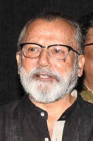 Man with glasses and a white beard smiles against a dark background, wearing a black shirt. The mood appears calm and content.