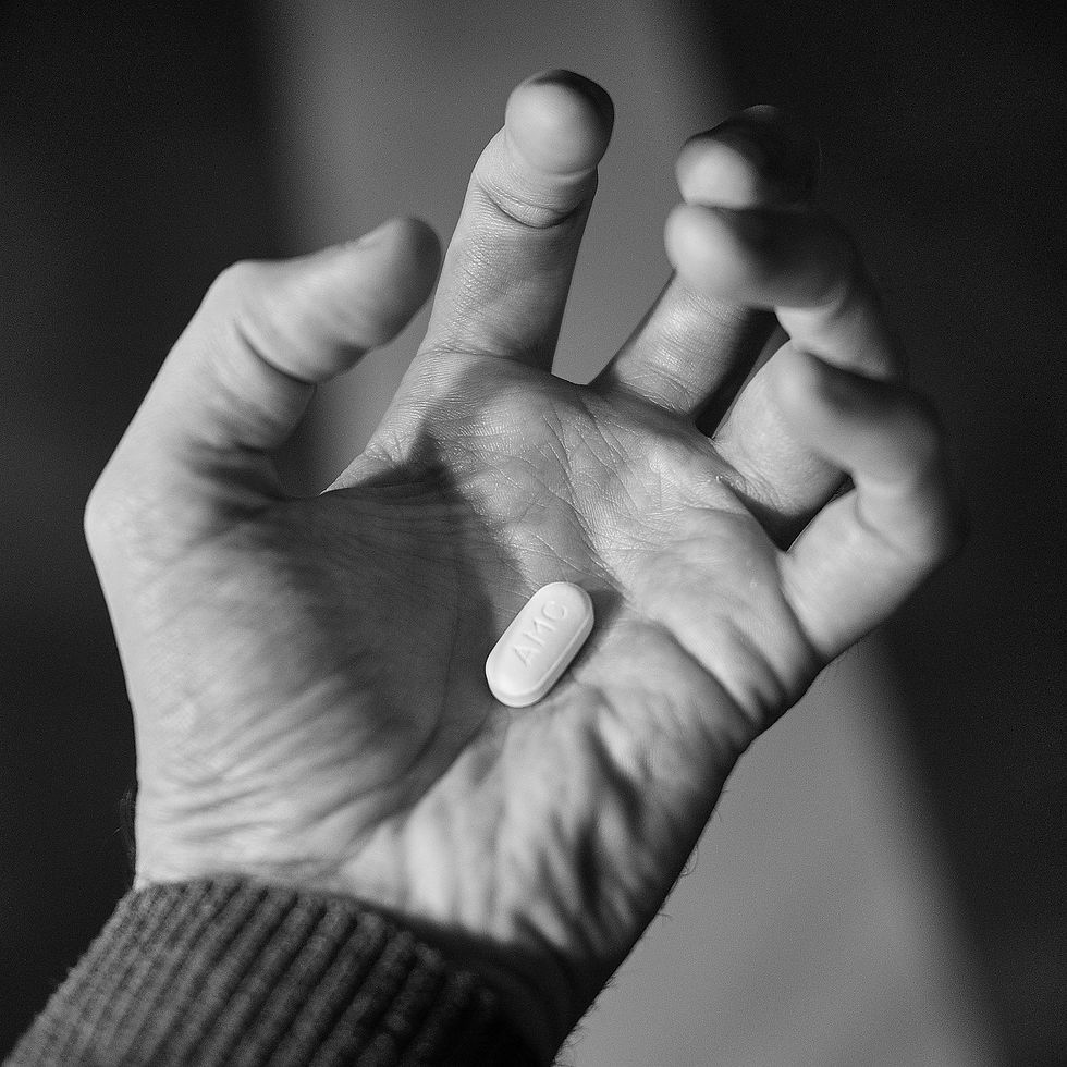 A hand holding a pill inscribed with "AMC," seen in black and white. The setting is dimly lit, conveying a contemplative mood.