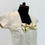 Thumbnail: A close-up side view of a beige linen dress displayed on a black mannequin against a plain white background. The dress features a square neckline and short sleeves with subtle seam detailing. The focal point is a decorative bow with a nature-inspired print, showcasing birds and foliage in earthy tones, positioned just below the neckline. The fabric’s texture and soft pleating are clearly visible, emphasizing the garment’s natural, handcrafted aesthetic.