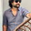 Thumbnail: Man in a grey linen shirt and dark sunglasses standing by a curtain, resting one arm on a wicker chair.