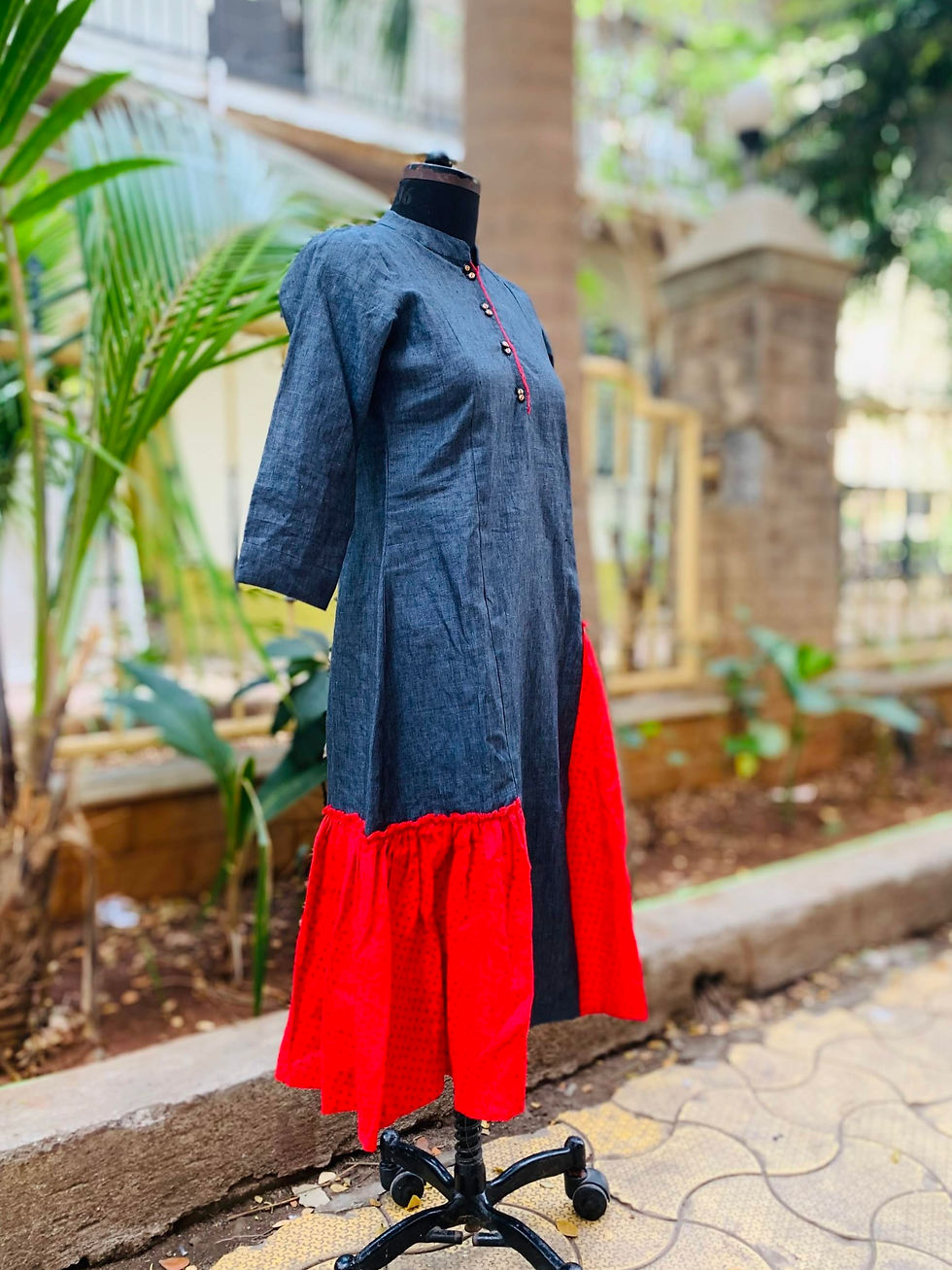 A mannequin displays a stylish, two-tone dress with a charcoal grey upper half and a vibrant red gathered hem. The kurta features three-quarter sleeves, a mandarin collar, and a front button placket accented with red piping and decorative buttons. It is set outdoors against a backdrop of tropical greenery and stone architecture