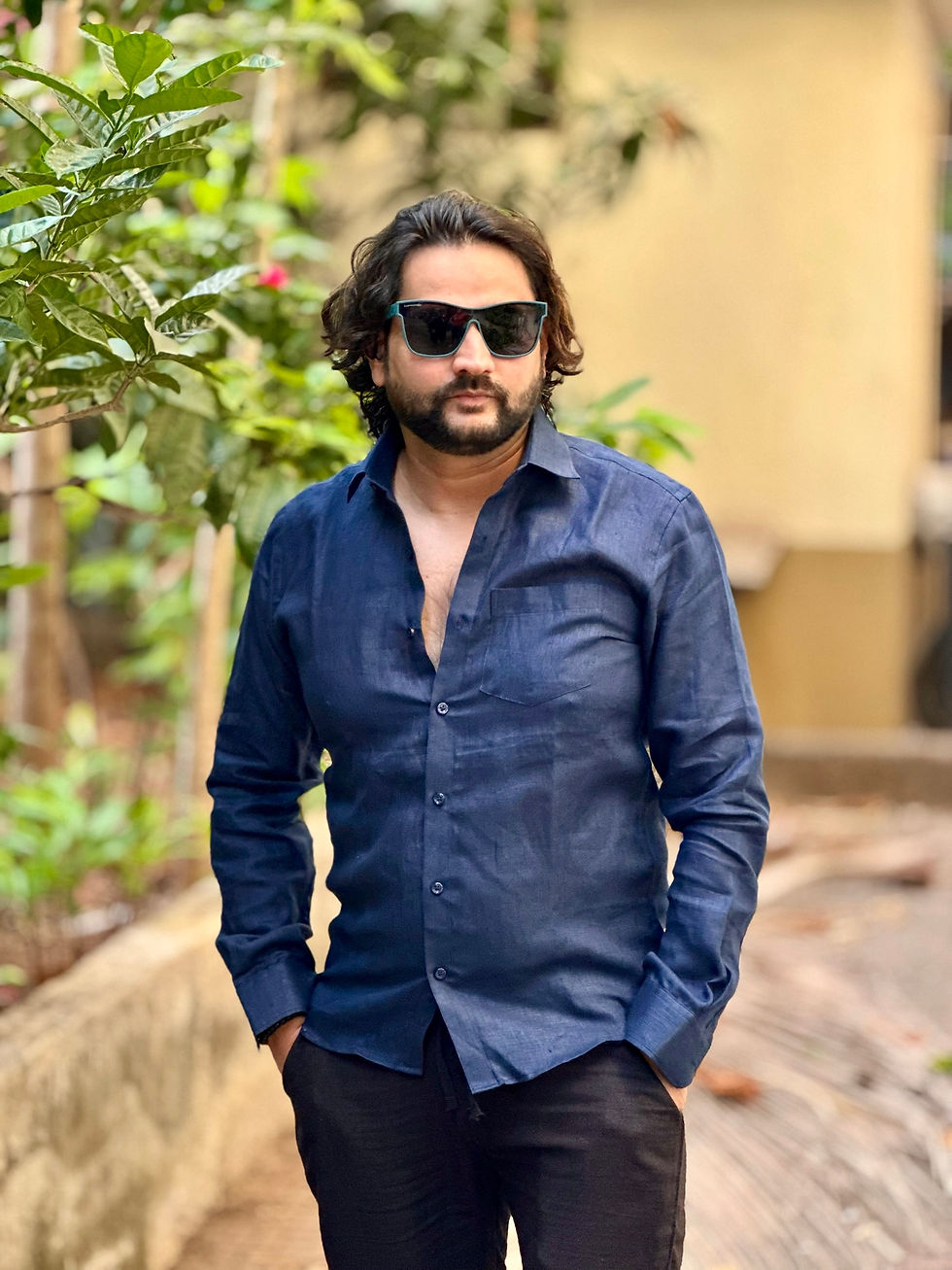 Thumbnail: Man in a Prussian blue linen shirt and black pants standing outdoors near green foliage, wearing black sunglasses.