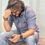 Thumbnail: Man in a blue shirt and jeans sitting indoors on a gray bench, smiling while looking at his phone, with one hand touching his