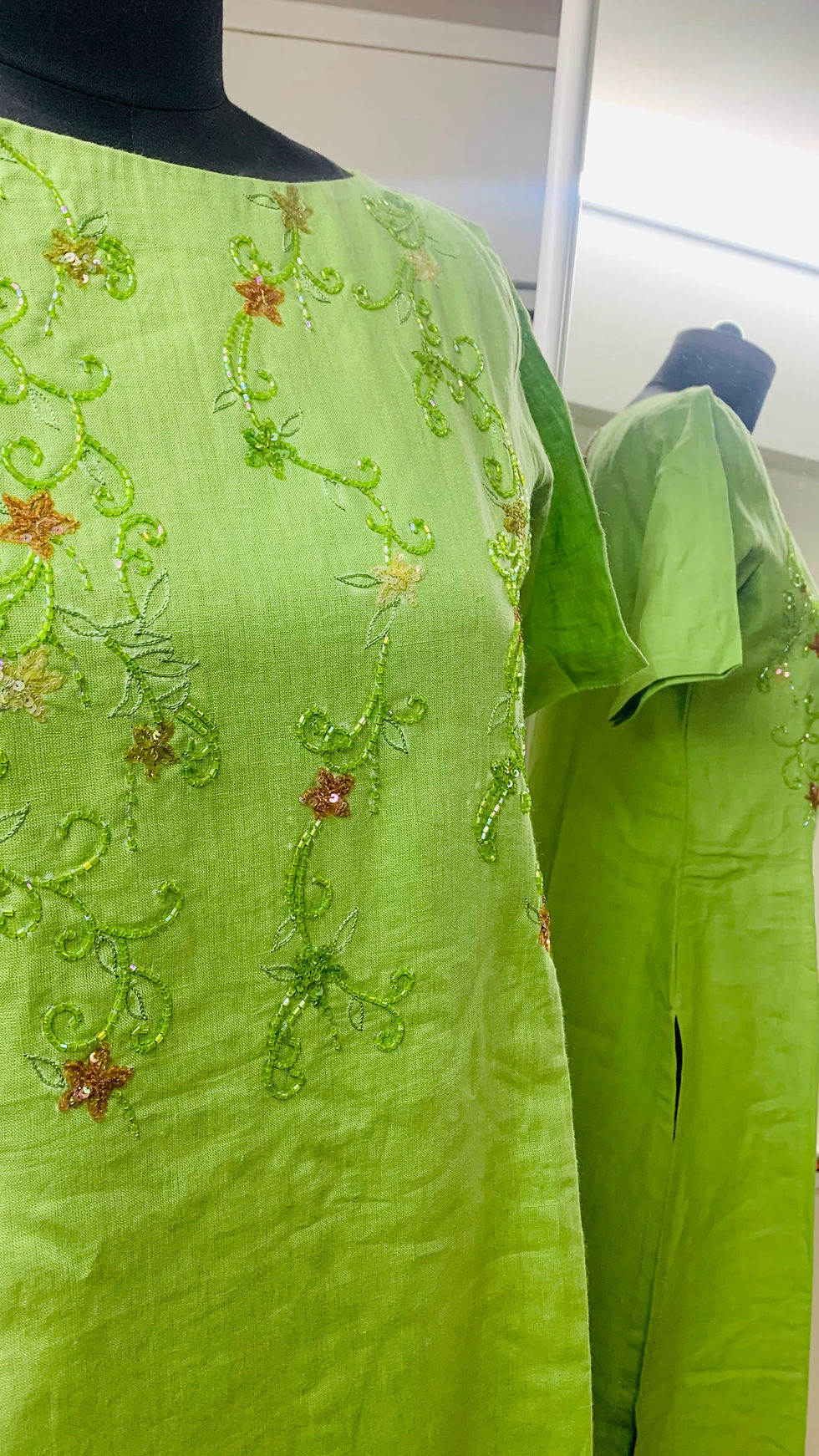A green pure linen kurta displayed on a black mannequin, featuring delicate hand embroidery with floral and vine motifs. The embroidery includes green, gold, and copper-toned threads and is embellished with sequins and beads, creating an elegant, shimmering effect. The kurta has short sleeves and a straight silhouette, with a slit visible at the side, reflected in the mirror behind.
