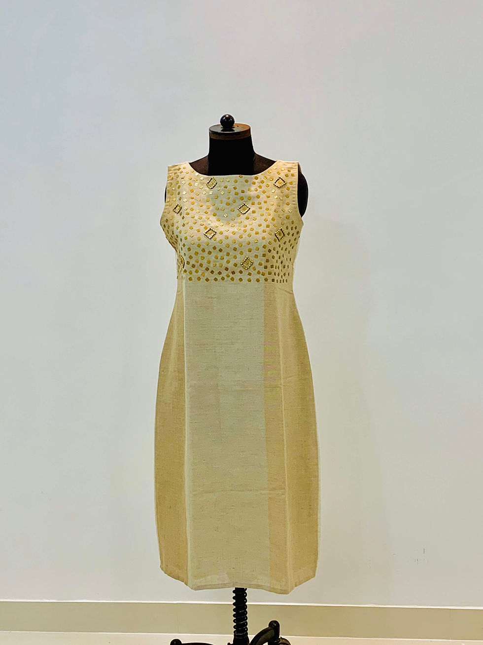 A beige sleeveless midi dress with gold embellishments on the bodice, displayed on a black mannequin in indoors .